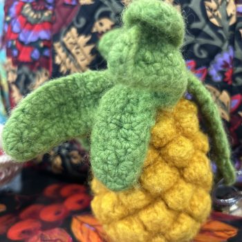 A crocheted pineapple with green leaves on a colorful fabric background.